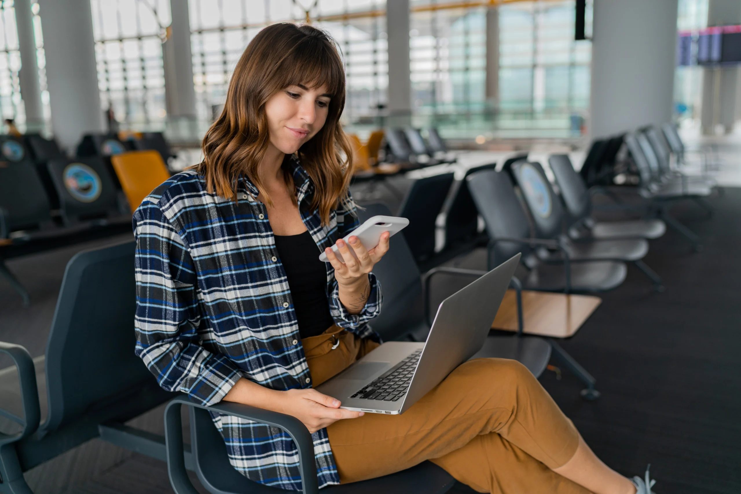 Young woman sitting in an airport terminal using her phone and laptop while waiting for her flight