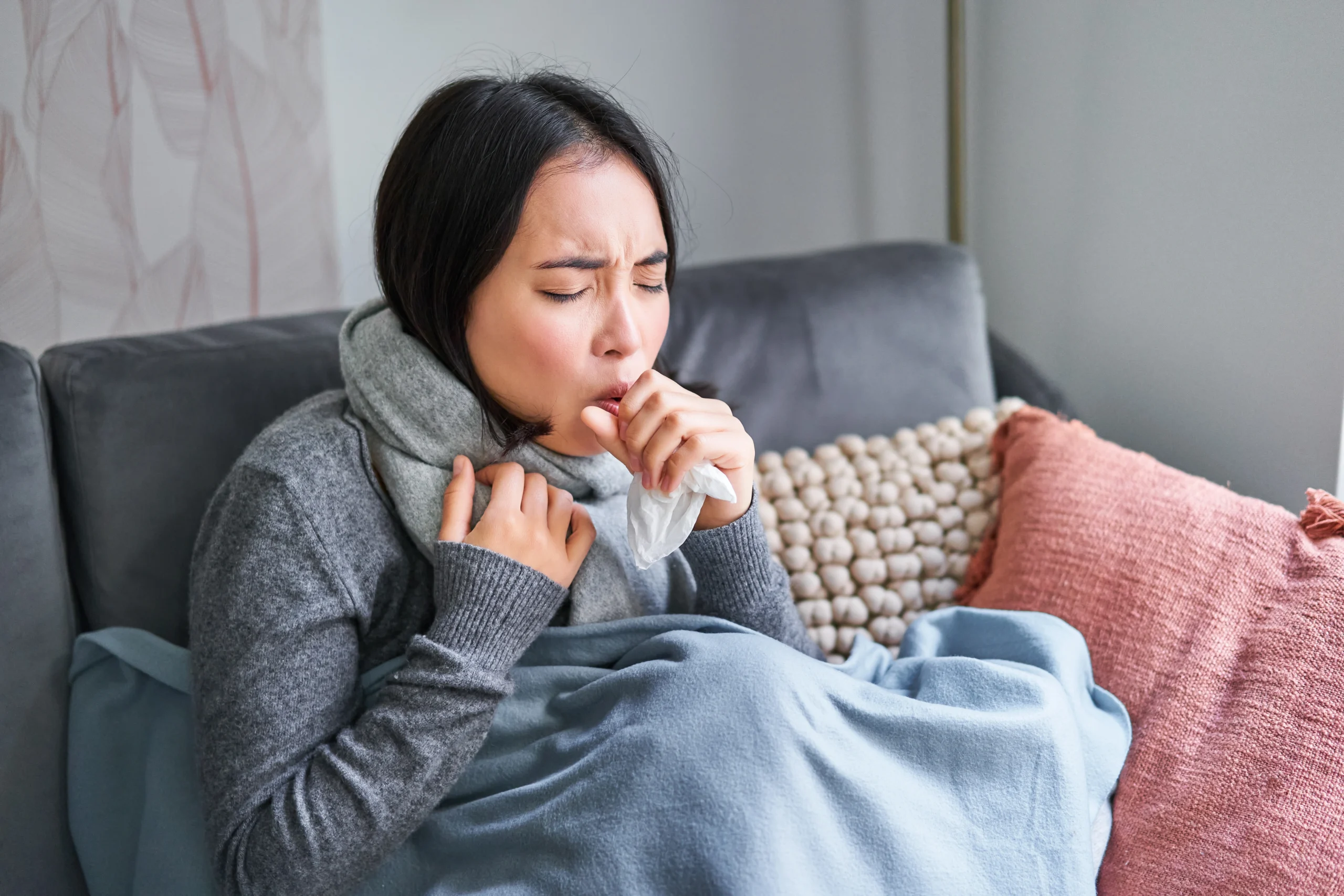 Young woman wrapped in a blanket and scarf, coughing into a tissue while resting on her couch with flu symptoms