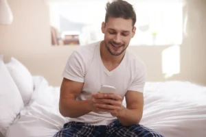 Man relaxing in bed at home using his phone to access telehealth care