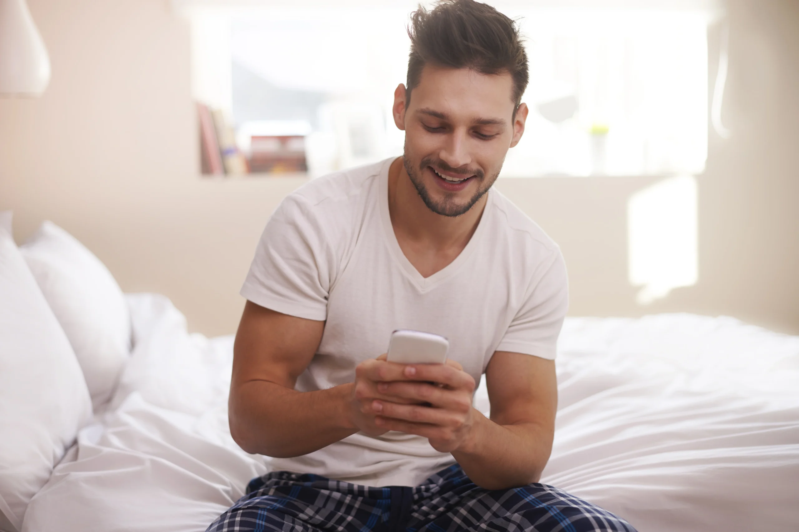Young man sitting on the edge of his bed in the morning, smiling while using his smartphone